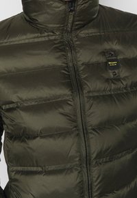 Olive green puffer jacket with a high collar, horizontal stitching, front zipper, and a logo tab near the chest. Smooth, shiny texture.