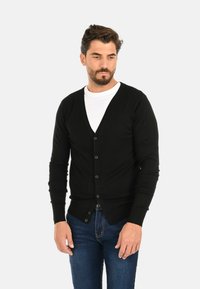 Cardigan nero con scollatura a V, chiusura con bottoni e polsini a costine. Indossato sopra una camicia bianca, abbinato a jeans blu. Materiale in maglia morbida.