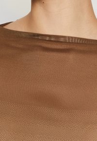 Zbliżenie na szyję i ramiona osoby noszącej brązowy, półprzezroczysty top z tkanej tkaniny o szerokim dekolcie.