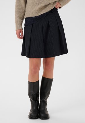 Personne portant un pull beige, une jupe noire plissée ornée de dentelle et des bottes noires montantes, debout sur un fond uni.