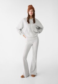 Šedá mikina s přední kapsou a šňůrkami, v kombinaci se šedými zvonovými kalhotami. Hnědý pletený kulich dodává kontrast. Měkká struktura na celém oděvu.