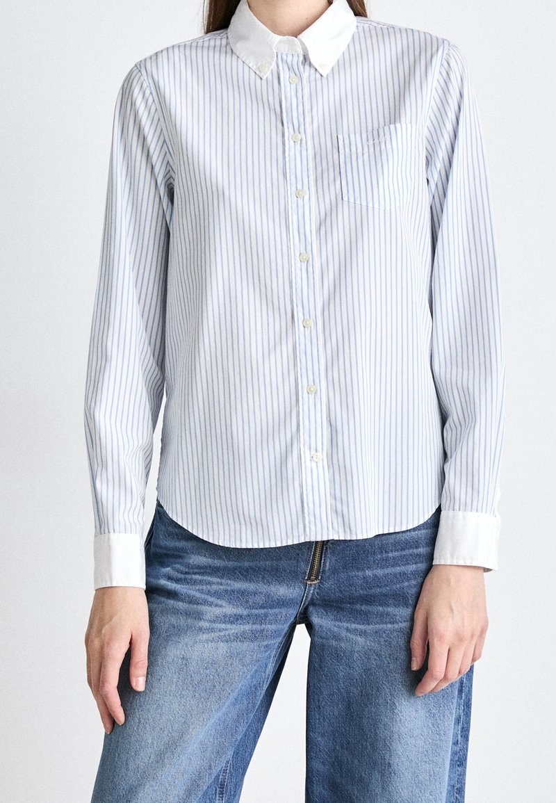 Personne portant une chemise à rayures verticales bleues et blanches à boutons, avec col et poignets blancs, associée à un jean bleu, debout.
