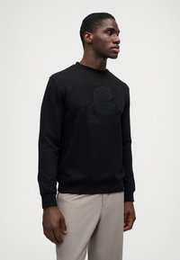 Μαύρη φλούτα με ανάγλυφη σιλουέτα μπροστά, στρογγυλή λαιμόκοψη και ραμμένα μανίκια. Φοριέται με ανοιχτόχρωμο παντελόνι.