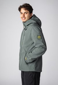 Lichtgroene jas met een capuchon, voorzien van een ritssluiting aan de voorkant en zakken. De linkermouw heeft een gele patch met de tekst "URBAN EDITION".