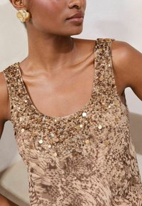 Beige sleeveless top with a sequined, textured neckline and a subtle animal print throughout. Gold circular earrings complement the look.