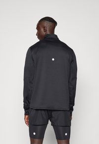 Svart atletisk jacka med hög krage, texturerat tyg och en vit cirkulär detalj på baksidan. Matchas med svarta shorts.