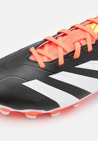 Botas de fútbol sintéticas negras con un acabado texturizado, cordones de color naranja brillante y acentos blancos. Diseñadas para ofrecer agarre y rendimiento en superficies de césped.
