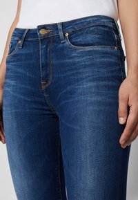 Jeans de un azul oscuro con una textura suave, tiro alto y herrajes dorados. Presenta un ligero desgaste y costuras de contraste a lo largo de las costuras.