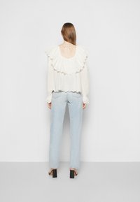 Blusa branca com pregas e textura rendada, costas abertas com um fecho de laço, combinada com jeans de perna reta azul claro e sapatos de salto bloco pretos.