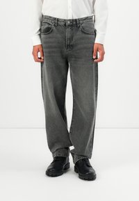 Man in losse grijze jeans, zwarte veterschoenen en een wit longsleeve shirt tegen een effen witte achtergrond.