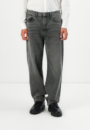 Man wearing loose gray jeans, black lace-up shoes, and a white long-sleeve shirt against a plain white background.