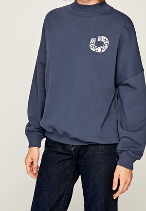 Person trägt ein marineblaues Sweatshirt mit langen Ärmeln, gerippten Bündchen und Kragen, mit einem kleinen weißen kreisförmigen Logo mit Text auf der Brust.