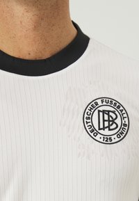 Valkoinen urheilupaita, jossa on pystysuorat raidat, musta ribbireuna kauluksessa ja pyöreä brodeerattu logo, jossa lukee "Deutscher Fussball-Bund 125."