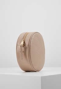 Runder, taupefarbener Kosmetikbeutel mit gestepptem Design, goldenem Reißverschluss und einer seitlichen Schlaufe für einfaches Handling. Glattes Kunstledermaterial.