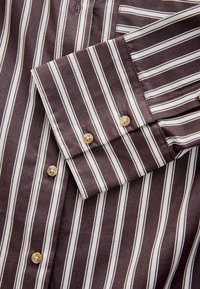 Camicia a righe marroni con strisce bianche, realizzata in tessuto morbido con una consistenza liscia. I polsini sono dotati di due bottoni marrone chiaro.
