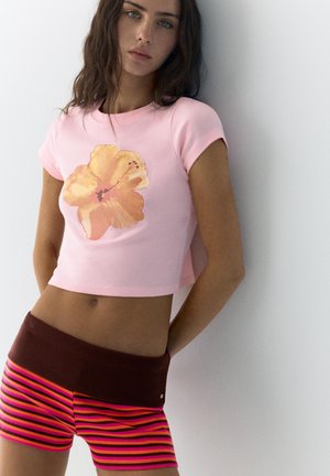 Tânără cu păr lung și închis la culoare, purtând un tricou roz cropped cu o floare portocalie mare și pantaloni scurți în dungi roz și maro.