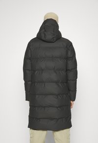 Lange zwarte pufferjas met een capuchon, voorzien van een gewatteerd ontwerp en een gladde textuur. De jas reikt tot halverwege de dij en heeft een relaxte pasvorm.