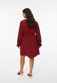 Robe bordeaux avec des manches ballons, taille cintrée, tissu texturé et accents brodés sur les manches. Associée à des mules à talons rouges assorties.