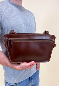 Bruine leren tas met een rechthoekige vorm, voorzien van een voorzak met rits, gestructureerd ontwerp en contrasterende witte stikselaccenten.