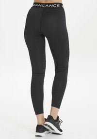 Schwarze Sportleggings aus dehnbarem Material mit einem hohen Taillenbund, der weiße Markenlogos und strukturierte Nähte aufweist. Kombiniert mit schwarzen Turnschuhen.