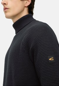 Dunkelblauer Rollkragenpullover mit strukturierter Strickmuster, ausgestattet mit einem gerippten Kragen und einem kleinen Logo-Patch am linken Ärmel.