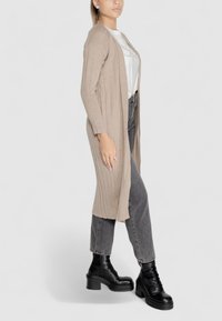 Cardigan beige a costine con maniche lunghe, indossato sopra una camicia bianca, abbinato a jeans grigi e stivali neri con tacco grosso.
