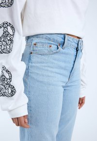 Jeans de mezclilla azul claro con cintura alta y cinco bolsillos, que presentan remaches de cobre y un acabado texturizado, combinados con una blusa blanca de manga larga.