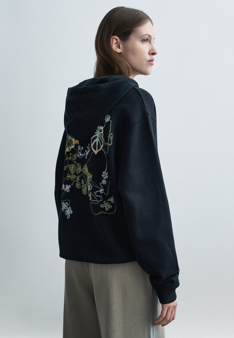 Γυναίκα με καστανά μαλλιά που φοράει μαύρο φούτερ με πολύχρωμη βοτανική κεντητική στο πίσω μέρος και μπεζ παντελόνι, σε πλαγιαστή όψη.