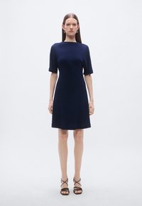 Rochie albastru-navy cu mâneci scurte și siluetă ușor în formă de A. Țesătură netedă cu decolteu rotund. Model purtând sandale negre cu barete.