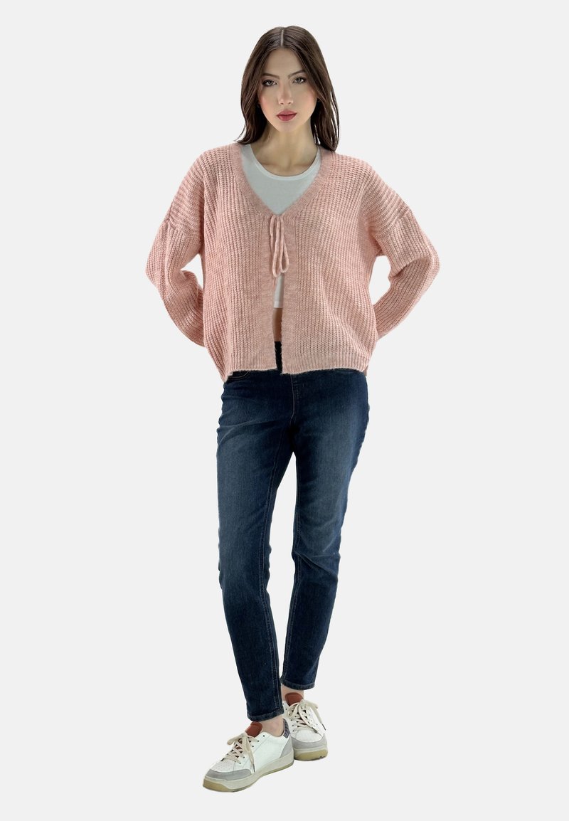 Hellblau gestrickte Cardigan mit V-Ausschnitt und Frontbindedetail, getragen über einem weißen Top, kombiniert mit dunkelblauen Jeans und lässigen Turnschuhen.