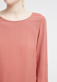 Rostfarbige Langarmbluse mit Rundhalsausschnitt und subtiler Schattierung. Verfügt über einen Knopfdetails an den Ärmeln zur Akzentuierung. Weicher, leichter Stoff.