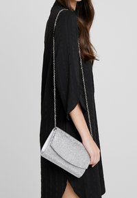 Pochette argentée ornée de sequins avec un rabat plié, une bandoulière chaîne, une forme rectangulaire et une texture lisse, tenue par une personne portant une robe noire.