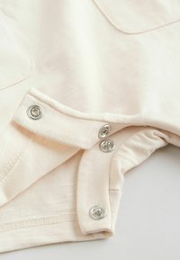 Nahaufnahme von beigem Stoff mit teils geöffneten silbernen Druckknöpfen an einem Kleidungsstückrand vor weißem Hintergrund.