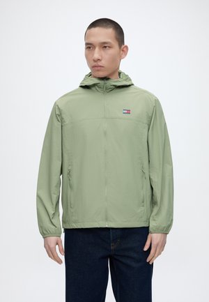 Mladý muž nosiaci svetlozelenú mikinu s kapucňou na zips s malým logom na hrudi a tmavomodré džínsy, stojaci pred jednofarebným pozadím.