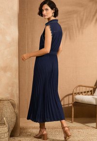 Marineblaues, plissiertes Maxikleid mit geknöpftem Kragen und gewelltem Saum. Verfügt über eine taillierte Taille und einen weiten Rock, kombiniert mit beigen Sandalen.