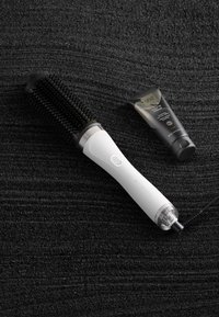 Sèche-cheveux brosse ronde avec un manche blanc, des poils noirs et un accent métallique. Comprend un tube de crème coiffante dans un emballage noir et or.