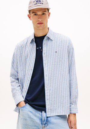 Nuori mies, jolla on päällään raidallinen sininen ja beige napillinen paita farkkutakin päällä, tumma laivastonsininen logo-T-paita, vaaleanpesut farkut ja vaaleanharmaa lippalakki, jossa on teksti.