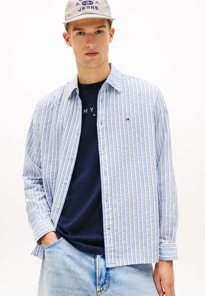 Νέος άνδρας που φοράει ριγέ πουκάμισο με κουμπιά σε μπλε και μπεζ αποχρώσεις πάνω από μπλε T-shirt με λογότυπο, τζιν ανοιχτού χρώματος και ανοιχτό γκρι καπέλο με κείμενο.