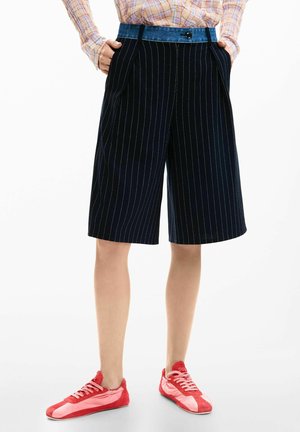 Személy sötétkék tűsarkú, térdig érő széles szárú rövidnadrágot visel, kockás hosszú ujjú inget betűrve, valamint piros fűzős sportcipőt rózsaszín részletekkel.