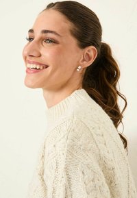 Weißer Kabelstrick-Pullover mit hohem Kragen, der detaillierte Muster aufweist. Das Model hat lange Haare, die zurückgebunden sind, und trägt silberne Creolen.