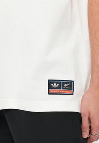 Balta kokvilnas T-krekls ar zilu un melnu plāksteri, uz kura ir Adidas logo un uzraksts "ORIGINALS FOR ORIGINALS" apakšmalā.
