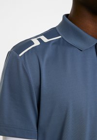 Kauluspaita tummansinisestä kankaasta, jossa on valkoinen diagonaalinen korostus olkapäällä ja sileä pinta.