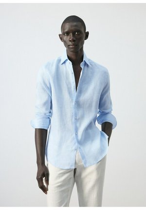 Uomo che indossa una camicia azzurro chiaro con maniche arrotolate e pantaloni chiari, in piedi con una mano in tasca.