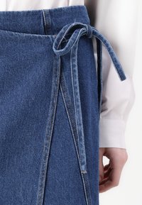 Denim omslagskjørt i blått, med knyting i livet, frynsete kanter og synlige sømmer, sammen med en hvit langermet skjorte.