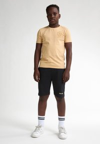 T-shirt en col rond beige avec logo sur la poitrine gauche, assorti à un short noir et des chaussures blanches. Pieds ornés de chaussettes rayées blanches et noires.