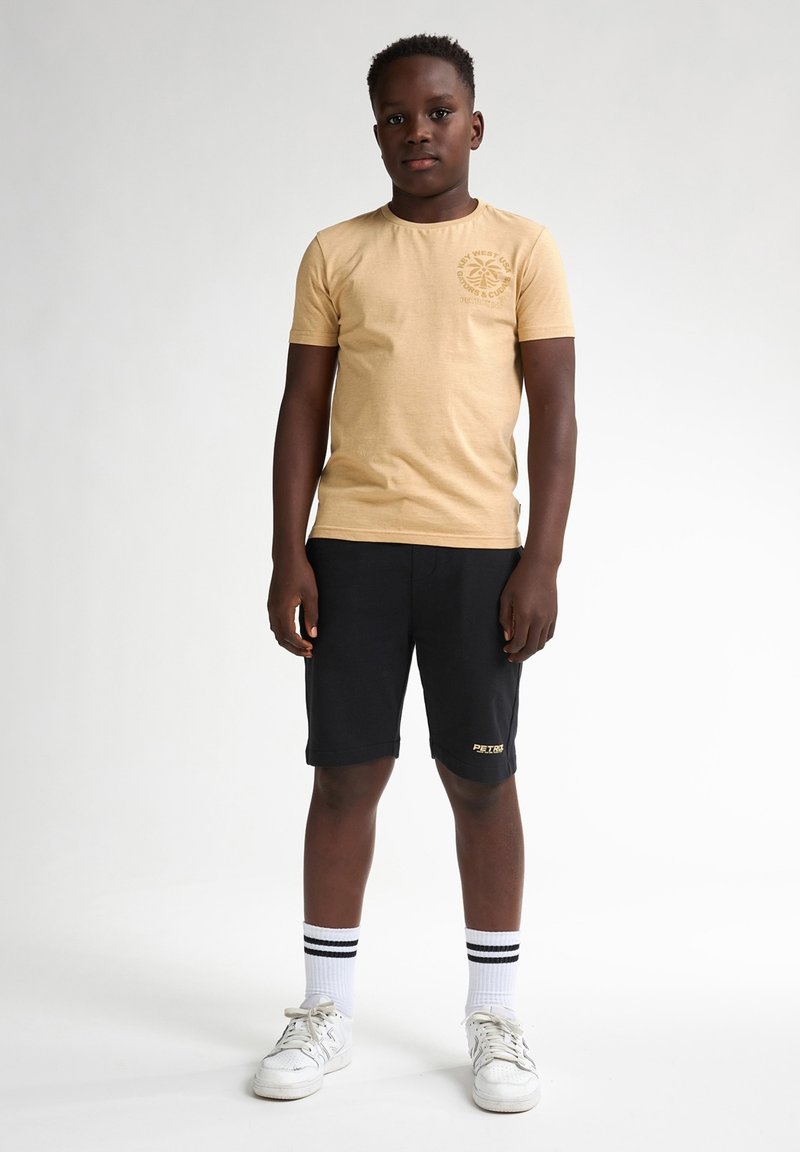 T-shirt en col rond beige avec logo sur la poitrine gauche, assorti à un short noir et des chaussures blanches. Pieds ornés de chaussettes rayées blanches et noires.