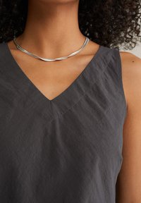 Top sans manches gris foncé avec un décolleté en V, fabriqué en tissu doux et légèrement texturé, avec des détails de couture contrastants. Collier en argent torsadé visible.