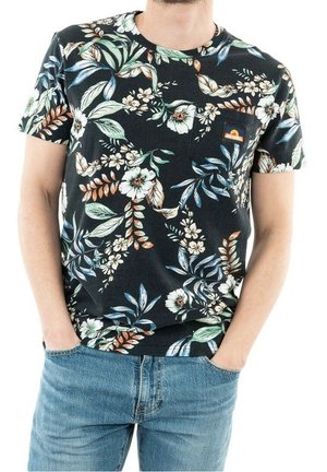 Mann trägt ein schwarzes Kurzarm-T-Shirt mit buntem Blumenmuster und blaue Jeans, die Hände in die Taschen gesteckt.