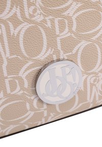 Beżowy, teksturowany materiał z wzorem białych liter. Na środku znajduje się okrągły srebrny emblemat z logo "JEANS".