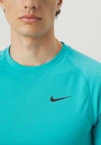 Türkises Nike Kurzarmshirt mit Rundhalsausschnitt. Mit einem schwarzen gestickten Swoosh-Logo auf der linken Brust. Glatte Stoffoberfläche.